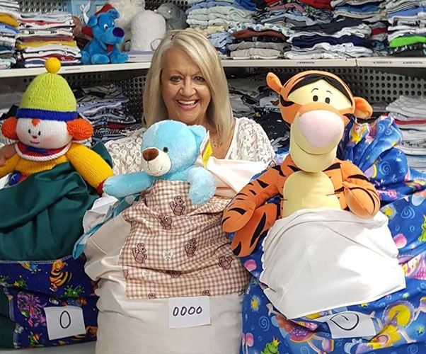 Lady with blond hair standing behind 3 very full large colorful handmade bags full of infant clothing. The bags are labelled with the sizes '0', 'oooo' and '1'. Each bag has a pocked with a large stuffed toy pocking out. Behind are full shelves neatly stacked with infant boy clothing.