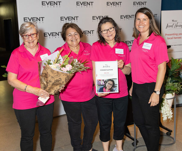 Four women volunteers in We Care pink tops and black pants. On the left the lady is holding a bouquet of flowers. Third from the left the lady is holding an award certificate with her photograph as the volunteer winner.