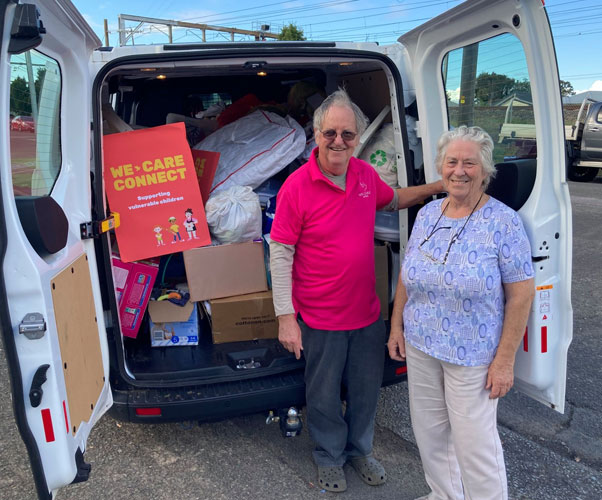 A man in a pink We Care tee-shirt and a woman with a lilac blouse and light gray trousers standing at the back of a van. The doors are open and the van is full of donated baby goods. The is a colorful sign saying We Care Connect